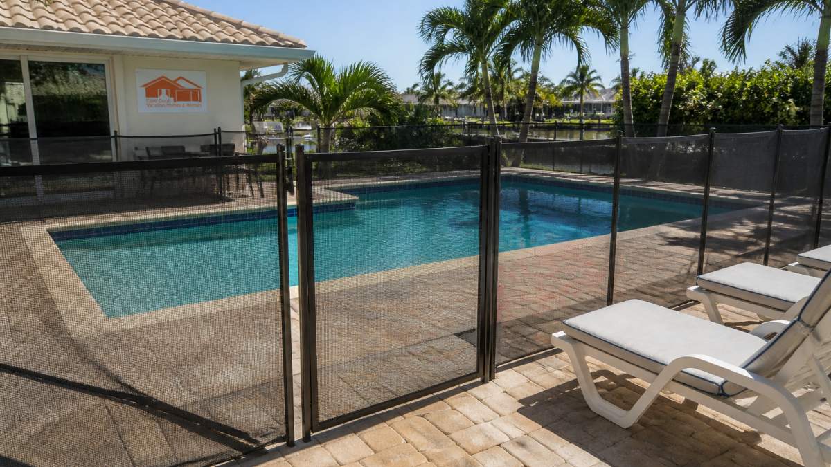 A sunny backyard pool at a Florida vacation villa with a screened safety fence and self-latching gate, palm trees in the background, and poolside lounge chairs under bright afternoon light